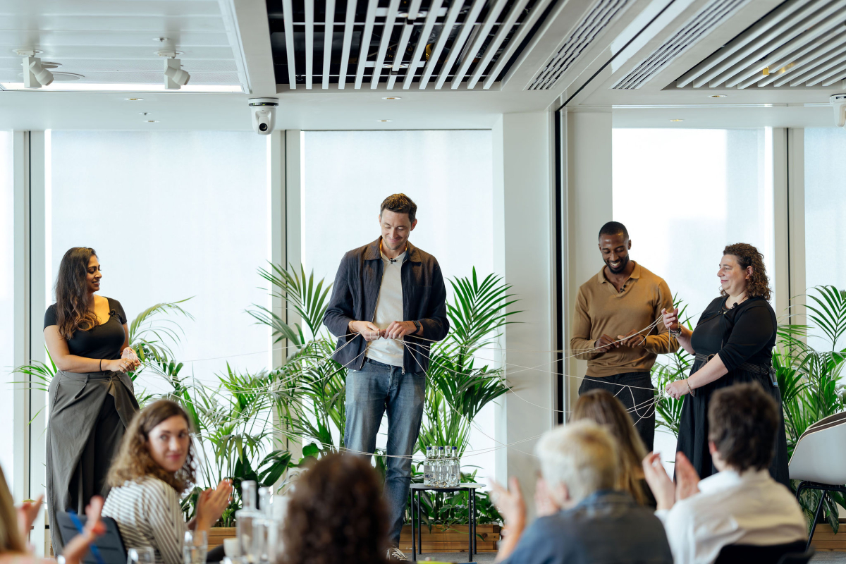 Four speakers stand at the front of the room during a session at Common Ground, holding a web of interconnected string to illustrate the connections between people, places and organisations in place-based giving. Delegates applaud in the foreground.
